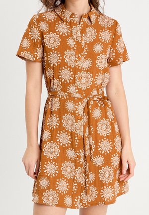 Femme portant une robe à manches courtes, boutonnée, de couleur rouille avec des motifs floraux blancs et une ceinture nouée à la taille.