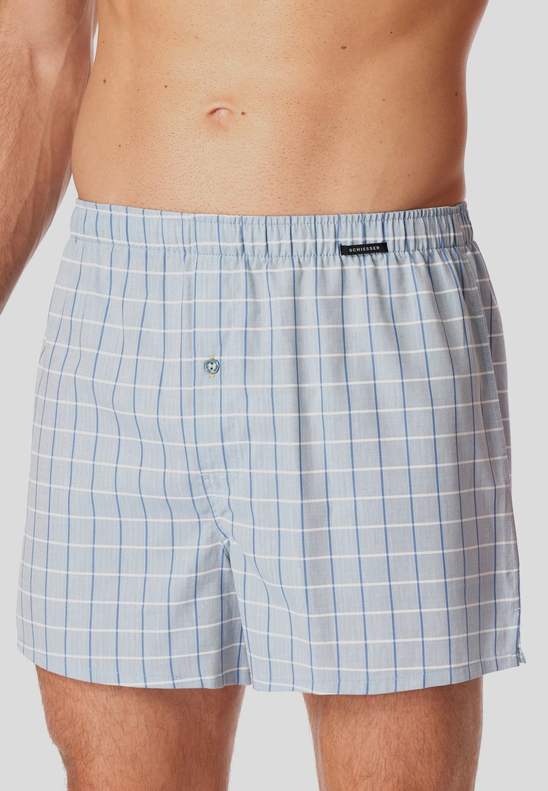 Boxer shorts de cuadros en azul claro y blanco para hombre, con una abertura de botón y cinturilla elástica, sobre el torso de un hombre de pie.