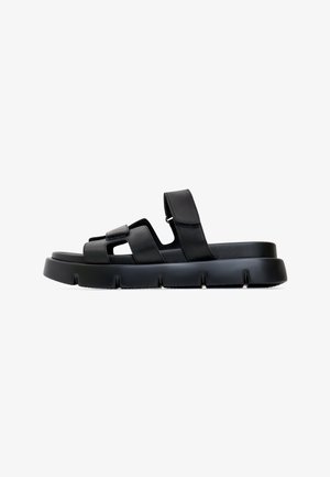 Sandalia negra con suela plana, correas ajustables y detalles recortados. Fabricada en material liso con un acabado texturizado en la suela.