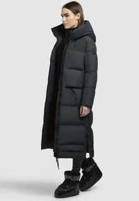 Lungo cappotto imbottito scuro con colletto alto, chiusura con zip e vita cinchata. Design a trama trapuntata con tasche laterali. Abbinato a stivali neri.