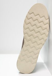 Suola di una scarpa taupe con un motivo a zigzag color crema, inclinata contro uno sfondo di muro e pavimento bianchi.