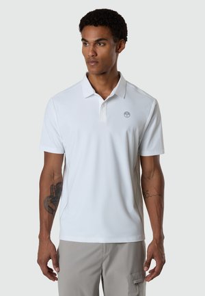 Mann mit kurzen lockigen Haaren trägt weißes kurzärmeliges Poloshirt und graue Cargohosen, zeigt Tätowierungen auf beiden Unterarmen, steht vor grauem Hintergrund.