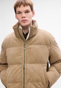 Tan ruskinds pufferjakke med høj krave, lynlås og sidelommer. Blød tekstur og kviltet design med kontrasterende panel.