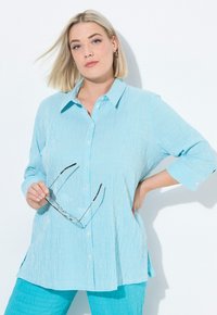 Camicia button-up a righe blu chiaro con colletto e maniche lunghe, indossata con pantaloni turchesi abbinati. Occhiali tenuti in una mano.
