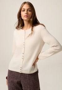 Cárdigan beige claro hecho de tela suave, con cuello redondo, mangas largas y botones decorativos en el frente.