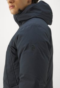 Navy blue insulated jacket with a hood, featuring a textured logo on the left sleeve and a front pocket for storage.