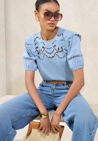 Helles blaues Blusenshirt mit Spitzenverzierungen, Puffärmeln und rundem Ausschnitt, kombiniert mit hochgeschnittenen Denim-Jeans und einem goldenen Armreif.