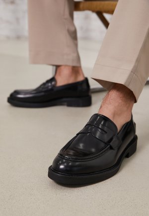 Black leather loafers with a smooth finish, featuring a decorative strap and contrast stitching, paired with beige trousers.