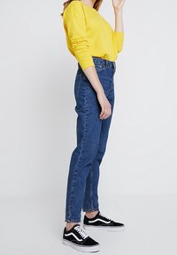 Felpa gialla, jeans blu a vita alta e sneaker nere con dettagli bianchi. I jeans sono a taglio dritto e arrivano fino alla caviglia.