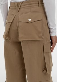 Beige cargobroek met een losse pasvorm, voorzien van meerdere grote zakken, een gladde textuur en zilverkleurige knopen op de achterzakken.