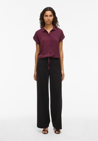 Woman wearing a short-sleeve burgundy button-up blouse with a front tie and black wide-leg pants with black open-toe heels.
