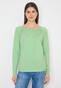 Femme portant un pull à manches longues vert clair avec un petit nœud sur le côté, associé à un jean bleu, debout devant un fond blanc uni.