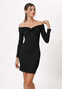 Abito nero off-shoulder con maniche lunghe, texture a costine, silhouette aderente e dettaglio a nodo sul busto. Tessuto liscio e elastico.