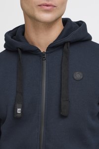 Mørkeblå zip-up hoodie med en sort lynlås, teksturerede snore og et rundt sort logo-patch på brystet. Blødt stof med en glat overflade.