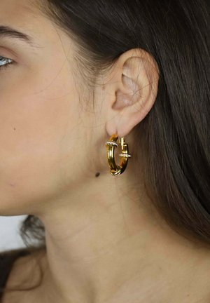 Złota kolczyk w kształcie koła z kręconym wzorem i małymi kryształowymi akcentami. Kolczyk ma błyszczące wykończenie i kompaktowy kształt.