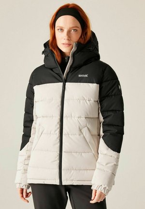 Femme aux cheveux roux portant un bandeau noir et une veste matelassée noir et blanc avec capuche, debout devant un fond uni.