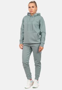 Set di felpa con cappuccio e pantaloni jogger in tessuto verde tenue, con tasca frontale, cordini e polsini elasticizzati. Indossati con sneakers bianche.