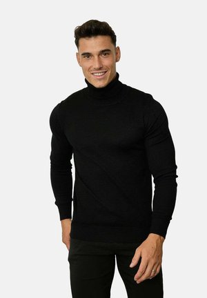 Pull à col roulé noir en tissu doux, avec des manches longues et des bords côtelés, porté sur un pantalon sombre.