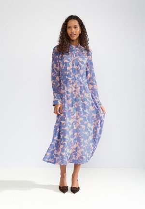 Vrouw met krullend haar, gekleed in een lange mouwen, knoopsluitende bloemenjurk in tinten blauw en roze, die de zoom van de jurk vasthoudt, staand op een witte vloer.