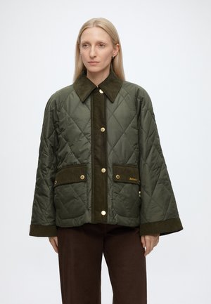 Vrouw met steil blond haar die een groene doorgestikt jas met gouden knopen en donkerbruine broek draagt tegen een effen witte achtergrond.