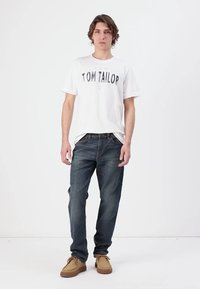 Valkoinen puuvillapaita, jossa on tummanharmaalla painettu "TOM TAILOR", yhdistetty tummansinisiin farkkuihin ja vaaleanruskeisiin nupukkinahkakenkiin. Seisova asento valkoista taustaa vasten.