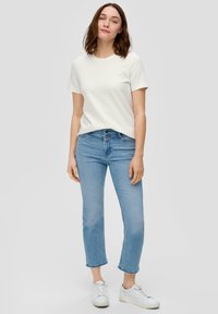 Witte t-shirt met korte mouwen gecombineerd met lichtblauwe straight-leg jeans. Model draagt witte sneakers en staat op een neutrale achtergrond.