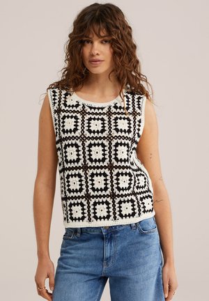 Femme aux longs cheveux bouclés portant un haut en crochet noir et blanc sans manches et un jean bleu, debout devant un fond neutre.