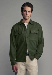 Grøn button-up jakke i et tekstureret stof, med to brystlommer, en krave og lange ærmer. Båret over en hvid skjorte.