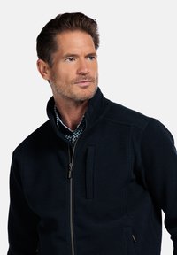 Marine blauw gestructureerde jas met een hoge kraag, ritssluiting aan de voorkant en zakken; gedragen over een gemusterd shirt, dat een moderne pasvorm en ontwerp laat zien.