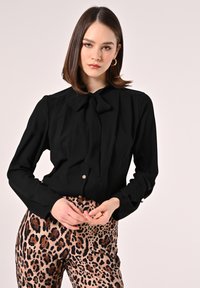 Blusa nera a maniche lunghe con colletto a fiocco e chiusura frontale con bottoni, abbinata a pantaloni aderenti beige con stampa leopardata.