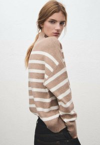 Maglione a righe beige e bianco in morbido tessuto testurizzato; presenta un collo alto e maniche ampie, abbinato a jeans scuri.