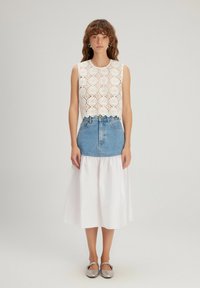 Haut sans manches en dentelle blanche avec des motifs circulaires ; associé à une jupe en denim bleue et une jupe inférieure blanche fluide. Des ballerines argentées complètent le look.