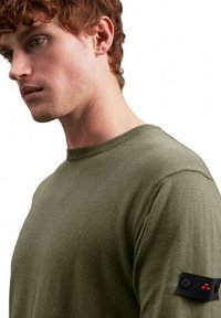 Olivgrüner Strickpullover mit Rundhalsausschnitt, mit einem schwarzen Stoffpatch auf dem Ärmel, der Knöpfe und rote Akzente aufweist.