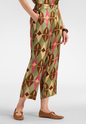 Femme portant un pantalon court taille haute avec des motifs géométriques verts, marron, roses et beiges et des chaussures plates marron, debout avec une main dans la poche.