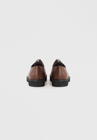 Brune lædersko med en glat finish, sorte gummisåler og snørebåndsfri design, der har afrundede kanter og minimalistiske detaljer.