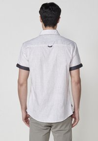 Camisa de manga corta en tejido gris claro con un acabado texturizado, que presenta detalles en gris oscuro en las mangas y un dobladillo redondeado.