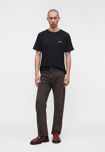 Άνδρας με μαύρο t-shirt A-COLD-WALL*, σκούρα καφέ παντελόνια με μαύρη ζώνη και μπορντό παπούτσια με κόκκινα κορδόνια, στέκεται μπροστά σε λευκό φόντο.