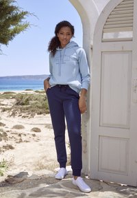 Helles blaues Hoodie mit Frontdruck, kombiniert mit dunkelblauen Sweatpants. Das Modell trägt weiße Sneakers und steht in einem Küstensetting nahe einer weißen Tür.