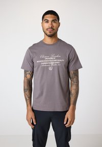 Graues Baumwoll-T-Shirt mit weißem Text, der Marken- und Kollektiondetails angibt, mit kurzen Ärmeln und lockerer Passform.