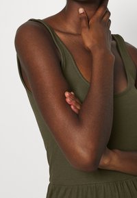 Oliwkowa, bezrękawnikowa bluzka z prążkowanego materiału, z okrągłym dekoltem. Zbliżenie na ramię i dłoń opartą na brodzie.