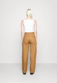 Žena s dlouhými blond vlasy, oblečená v bílém bezramínkovém topu a hnědých širokých kalhotách s vysokým pasem, otočená zády k jednobarevnému pozadí.