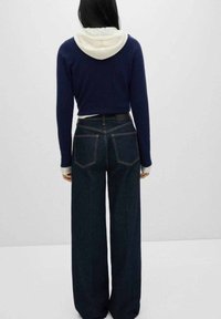 Marineblauwe trui met witte capuchon, gecombineerd met hoog-waisted wijde spijkerbroek van donkerdenim met contrasterende stiksels en twee achterzakken.