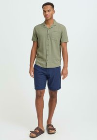 Chemise verte à manches courtes avec des boutons et une poche poitrine, associée à un short bleu marine et des sandales marron dotées de deux brides réglables.