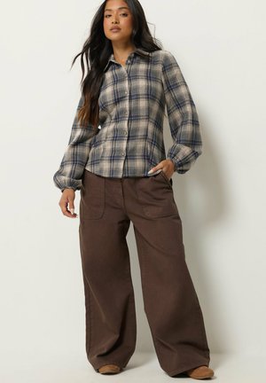Femme aux longs cheveux foncés porte une chemise à carreaux bleue et grise boutonnée et un pantalon marron ample, debout sur un fond blanc cassé.