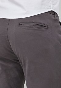 Ciemnoszare spodnie wykonane z gładkiej tkaniny, wyposażone w tylną kieszeń z poziomą klapką i subtelne detale przeszyć.