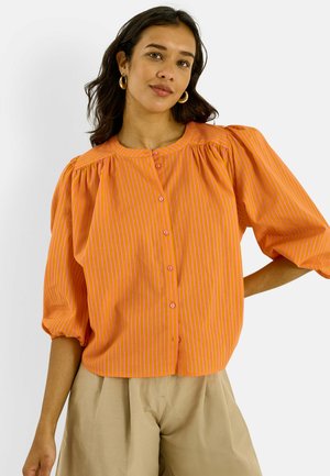 Femme portant un chemisier rayé orange et rose avec des manches bouffantes et un pantalon plissé beige, posant devant un fond blanc.