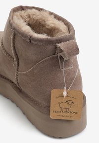 Brązowy zamszowy botek za kostkę z miękkim futrzanym wyściółką, zaokrąglonym noskiem i pętelką do ściągania. Zawiera metkę z napisem "VERO MONTONE" oraz logo owcy.