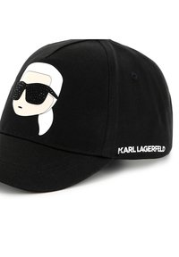 Melna beisbola cepure ar grafiku, kurā attēlots sejas attēls ar baltām matu šķipsnām un melnām saulessargu brillēm, ar uzrakstu "KARL LAGERFELD" izšūtu baltā krāsā.
