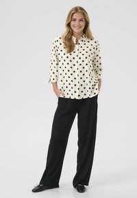 Blusa a pois color crema con punti neri, apertura con bottoni nella parte anteriore, maniche con polsino. Abbinata a pantaloni neri a gamba larga e scarpe basse.