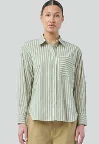 Modellen bærer en grøn og hvid stribet skjorte med knap, der har en brystlomme, og er iført beige bukser, mod en neutral grå baggrund.